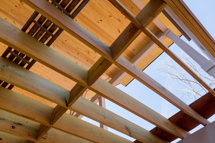 View from below of wooden beams and panels forming a roof structure, showing blue sky through the gaps.