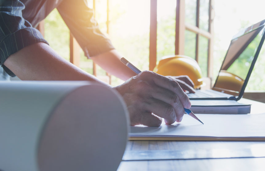 Person writing on paper at a desk with architectural plans, two yellow hard hats, and an open laptop nearby.