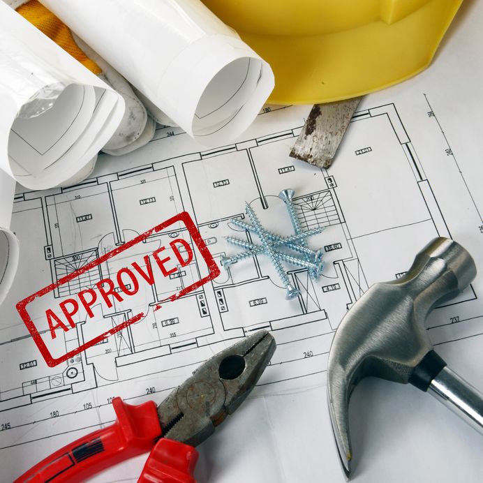 Construction tools on an approved blueprint with a rubber stamp mark, hammer, pliers, screws, and a hard hat.