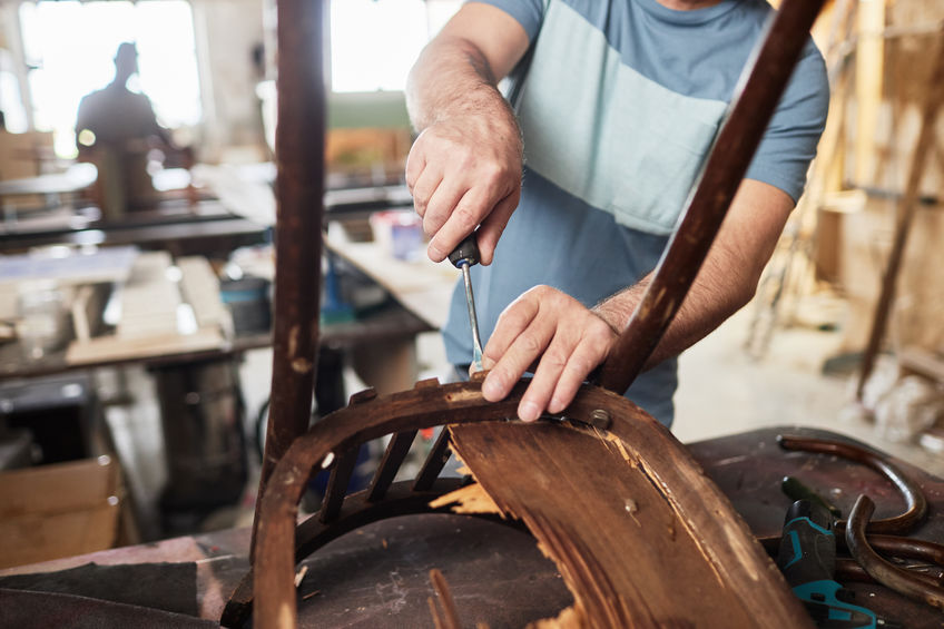 Person repairing a wooden chair, using a screwdriver, in a workshop setting with tools and materials in the background.