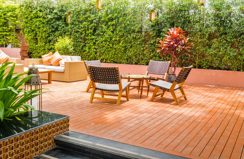 Outdoor patio with wooden flooring, cushioned seating, a small table, lush greenery, and a red-leafed plant.