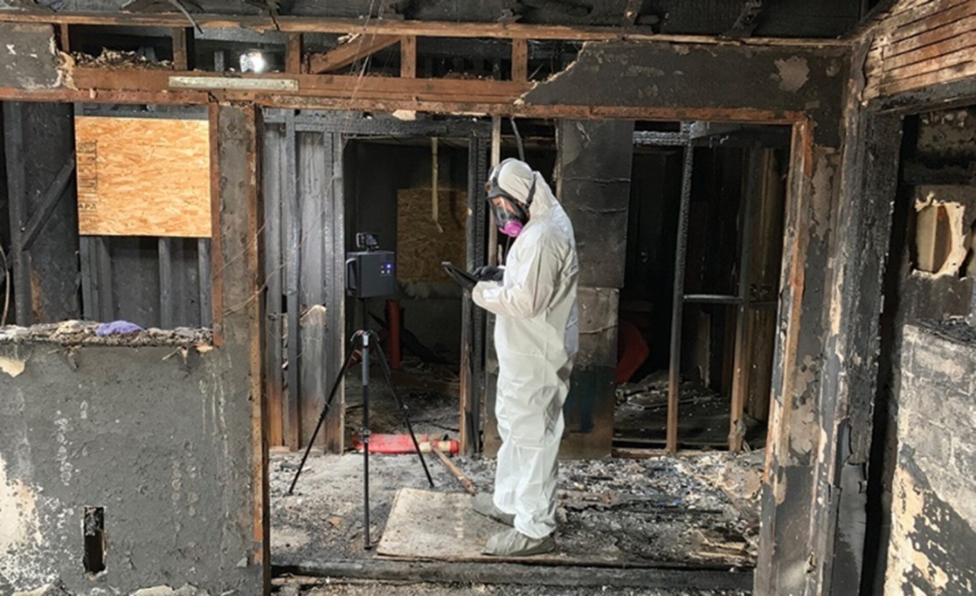 Person in protective gear examines fire-damaged room with charred walls, using equipment on a tripod.