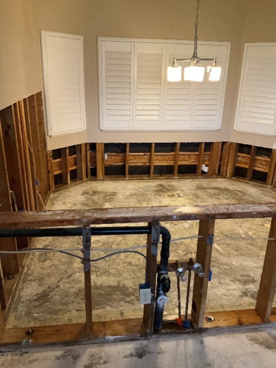 Room under renovation with exposed wooden framing, plumbing, and no flooring. Overhead light fixture and shuttered windows visible.