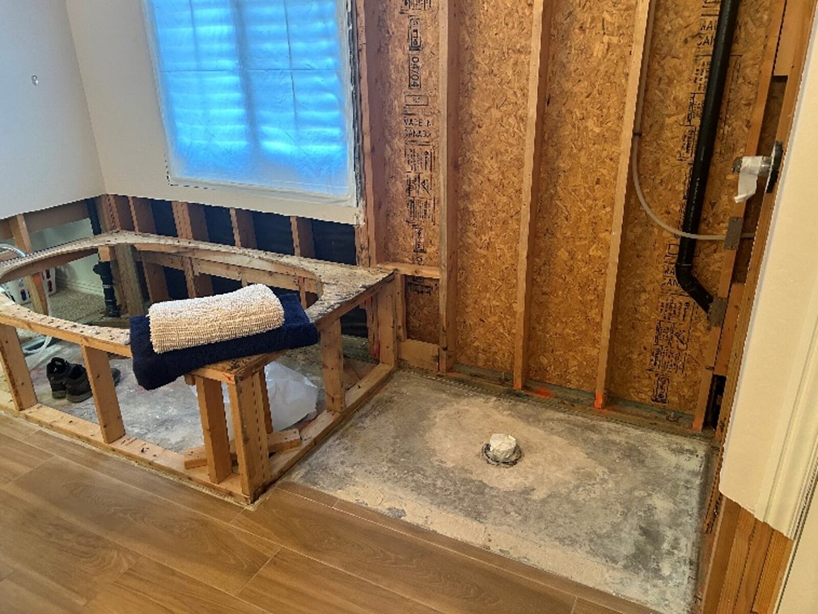 Under-construction room with exposed wooden studs and plumbing, a stack of towels on the framing, and wood flooring installed.