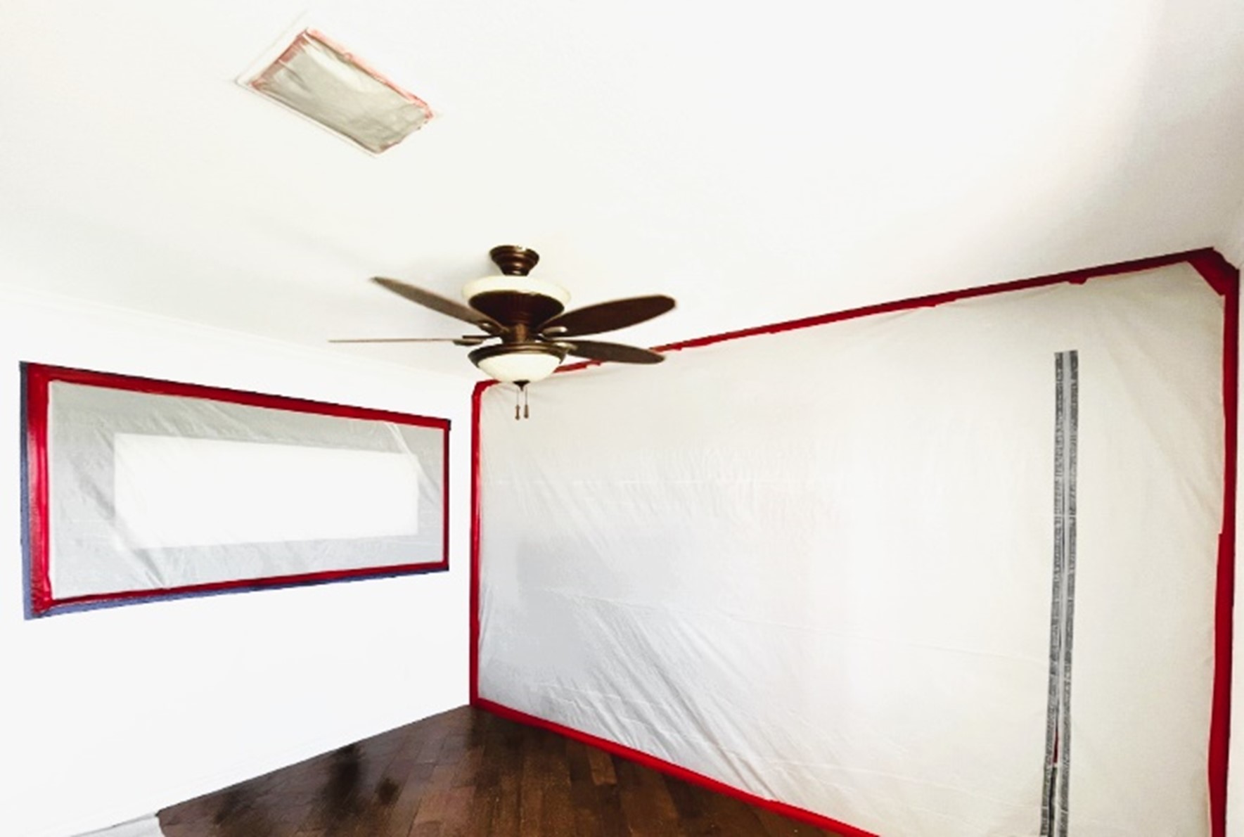 Room with hardwood floor, white walls, red tape, plastic sheeting on window, ceiling fan, and sealed air vent.