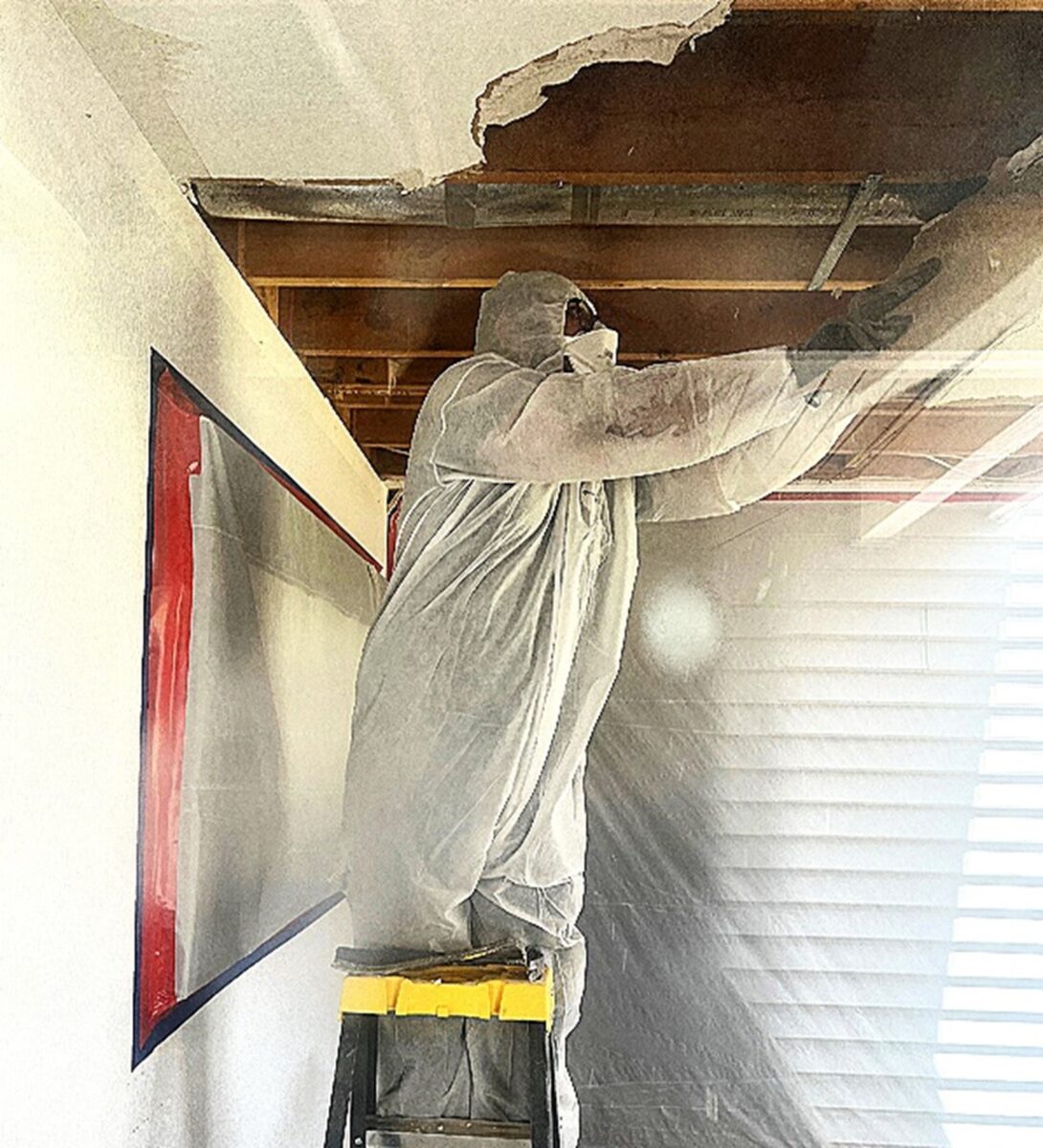 Person in protective gear repairing a damaged ceiling while standing on a ladder.