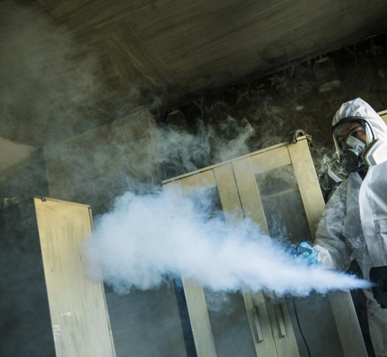 Person in protective suit fumigating a room with smoke, standing near a wardrobe in a dimly lit space.