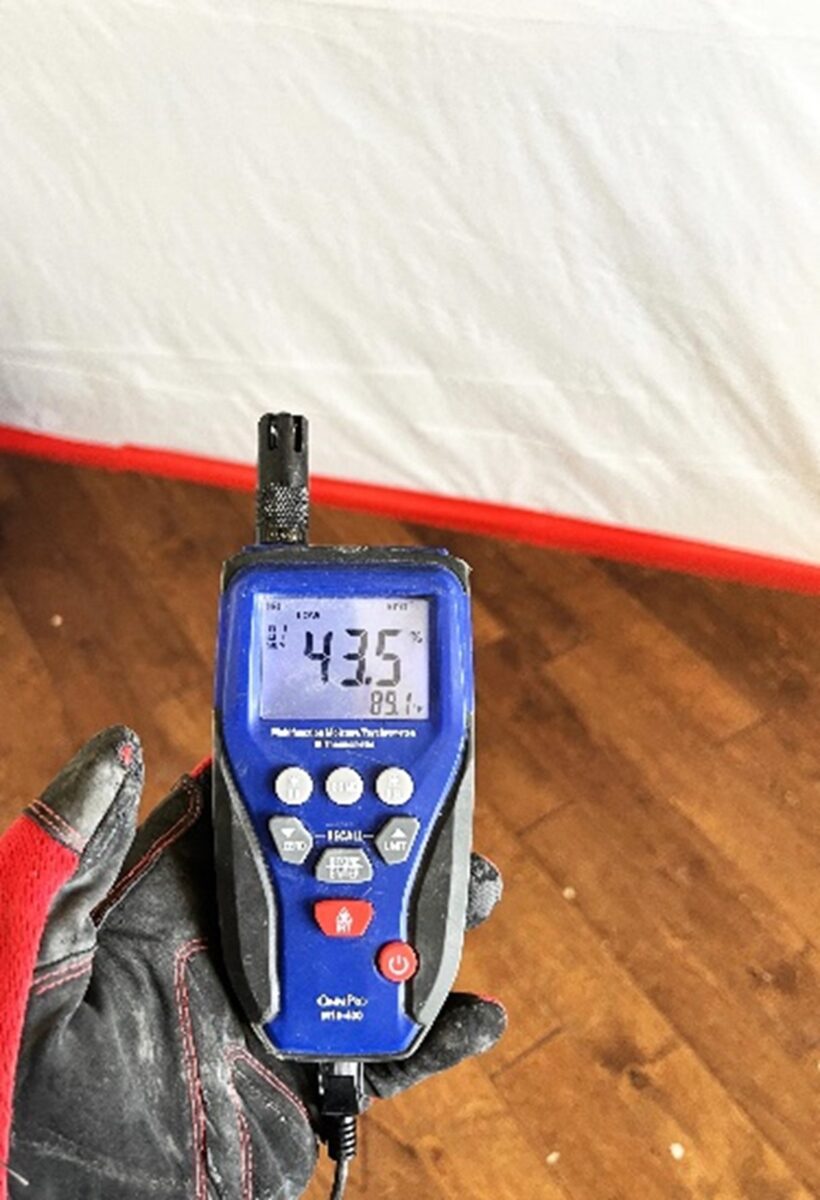 Hand holding a digital device displaying 43.5% humidity and 89.1°F. Device has buttons and a probe, background shows a floor.