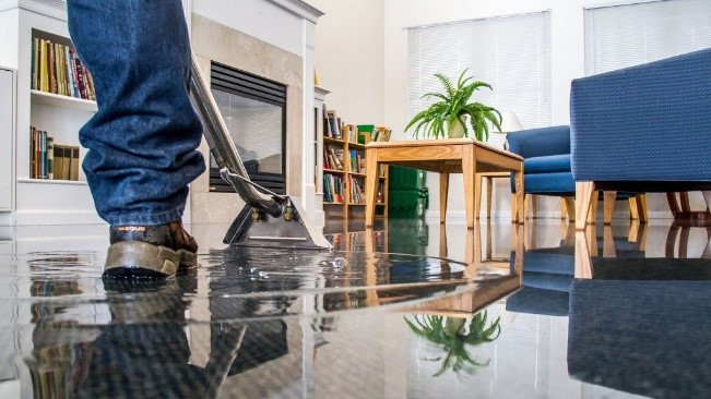 A person uses a vacuum to remove water from a flooded living room, with a blue sofa and bookshelves in the background.