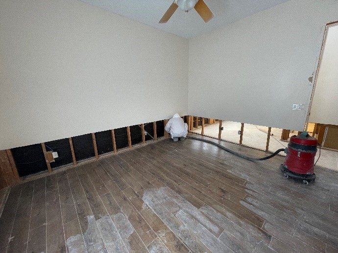 Person in protective suit working on exposed wall studs in a room with wooden flooring. A shop vacuum is nearby.